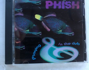 May include: A CD case with a black background and a colorful image of a fish. The text "Phish" is in purple and the text "Playing in the club" is in blue.