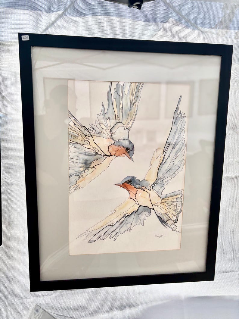 K&ouml;nnte beinhalten: Ein gerahmtes Aquarellbild, das zwei fliegende Schwalben zeigt. Die V&ouml;gel sind mit sanften Waschungen in Blau-, Grau- und Pfirsicht&ouml;nen dargestellt, die durch feine schwarze Tintenumrisse definiert sind. Das Kunstwerk ist in einem schwarzen Rahmen.