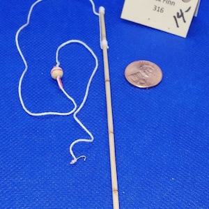 Może przedstawiać: Drewniana wędka z białą żyłką i przymocowaną czerwoną i białą kulką. Wędka ma około 14 cali długości. Wędka leży na niebieskiej powierzchni z groszem jako skalą.