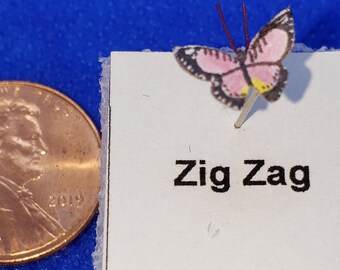 Peut inclure: Un petit carré blanc avec un papillon rose sur une épingle et le texte "Zig Zag" imprimé dessus. Le carré est posé sur une surface blanche texturée.