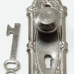 May include: Silver door knob and keyhole with ornate detailing. The knob is round and has a decorative design. The keyhole is oval shaped. The key is a simple design with a round head.