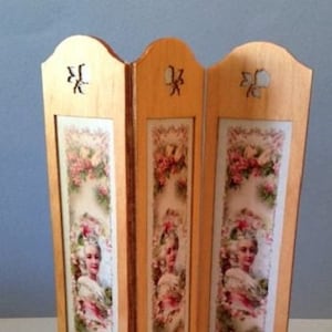 May include: A three-panel room divider made of wood with a light wood finish. Each panel features a floral design with a woman's portrait in the center. The divider is approximately 12 inches tall.