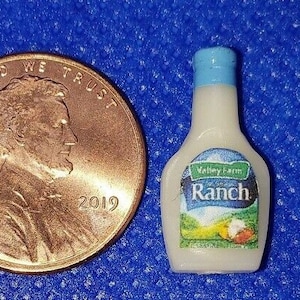 May include: Miniature Valley Farm Ranch dressing bottle with a blue cap, next to a 2019 penny. The label on the bottle features a landscape scene and the word "Ranch". The penny shows the profile of Abraham Lincoln.