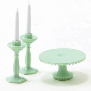 Puede incluir: Un soporte para pasteles en miniatura verde menta y dos candelabros a juego con velas blancas. El soporte para pasteles tiene un borde festoneado y una base de pedestal. Los candelabros tienen un diseño clásico.