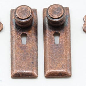 May include: Two rectangular copper-toned metal escutcheon plates, each featuring a circular knob at the top and a keyhole opening, accompanied by two matching skeleton keys. The metal hardware has a weathered, textured finish throughout.
