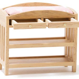 May include: A miniature wooden changing table with a light oak finish, featuring two small pull-out drawers with gold-toned knobs, two open storage shelves below, and a soft pink padded top surface with a subtle white pattern for dollhouse use.