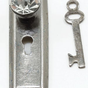 May include: A rectangular silver-toned metal escutcheon plate featuring a clear, faceted crystal knob at the top and a central keyhole opening. Beside the plate lies a matching silver-toned metal skeleton key with a rounded bow and bit.