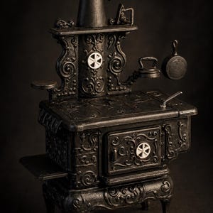 May include: A detailed miniature black cast iron stove with ornate scrollwork and white accents. The stove features a chimney, a small kettle, and a frying pan. The stove is a vintage style, perfect for dollhouses or miniature collections.
