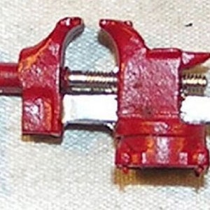 Peut inclure: Un petit étau en métal rouge avec une poignée argentée. L'étau est ouvert et prêt à serrer.