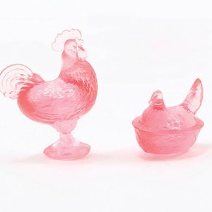 May include: Two pink glass figurines: a rooster standing upright and a hen sitting in a nest. The rooster has a detailed comb and tail feathers. The hen is in a nest-like bowl. Both are translucent.