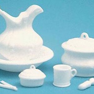 May include: A white ceramic miniature wash set consisting of a pitcher and basin, a lidded chamber pot, a small lidded container, a mug, a toothbrush, and two thin sticks. The pieces are solid white with a smooth finish and simple molded details.