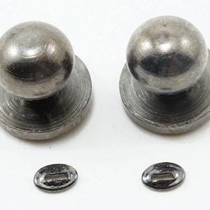 May include: Two silver metal knobs with a round top and a flat base. The knobs are on a white surface. Two small silver metal plates are also on the surface.