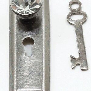May include: A rectangular silver-toned metal escutcheon plate featuring a clear, faceted crystal knob at the top and a central keyhole opening. Beside the plate lies a matching silver-toned metal skeleton key with a rounded bow and bit.