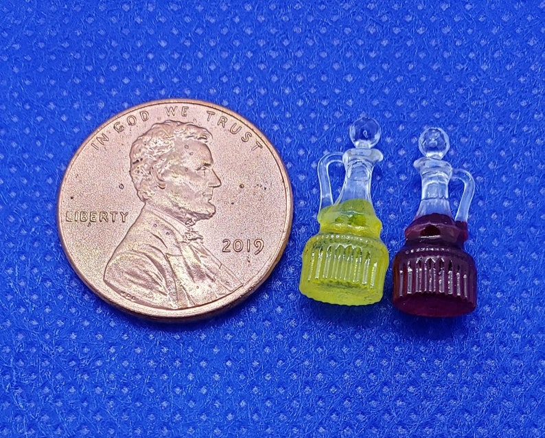 May include: Two miniature glass bottles with stoppers. The bottle on the left is filled with a yellow liquid and the bottle on the right is filled with a purple liquid. The bottles are sitting on a blue surface.