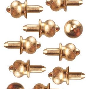 May include: A collection of twelve gold-colored metal studs, each featuring a rounded, bulbous head and a smooth, cylindrical post. The studs are arranged in a grid pattern, showing both side profiles and top-down views of the rounded heads.
