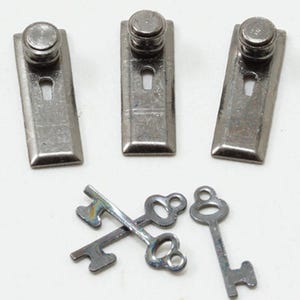 May include: Three rectangular silver-toned metal escutcheon plates, each featuring a small circular knob at the top and a keyhole cutout. Below the plates are three small silver-toned metal keys, with two of the keys positioned in a crossed arrangement.