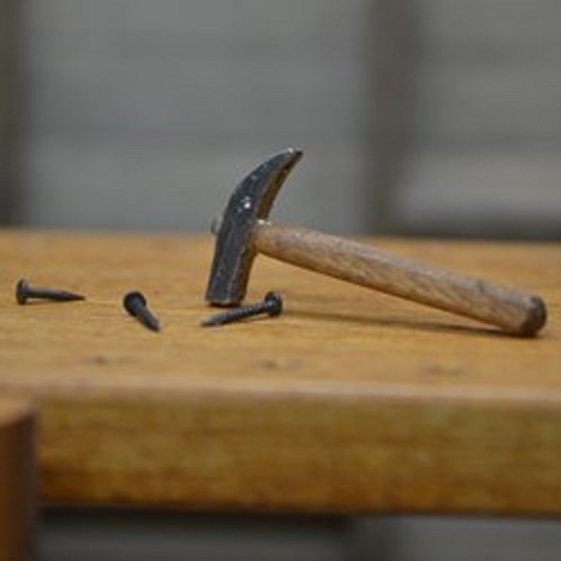 May include: A claw hammer with a wooden handle and a metal head rests on a wooden surface, alongside three black nails. The hammer is positioned as if it has just been used. The scene suggests a carpentry or DIY project.