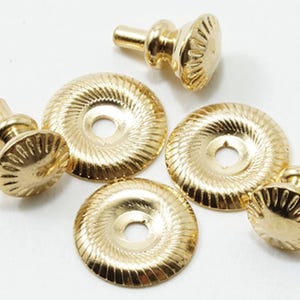 May include: A set of three gold-toned metal studs and two matching circular washers. Each stud features a rounded, textured top with radiating lines, while the flat washers have a corresponding grooved pattern around their outer edges.