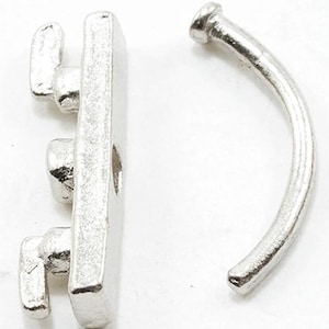 Puede incluir: Un cierre de joyería de metal plateado compuesto por dos piezas distintas. Una pieza es una barra rectangular con tres pequeños ganchos que sobresalen, y la otra pieza es una varilla curva semicircular con una tapa de extremo redondeada.