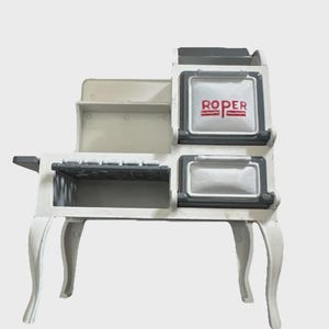 May include: A white toy stove with gray accents and curved legs. The stove has two oven doors and a shelf. The word "ROPER" is printed in red on the top oven door.