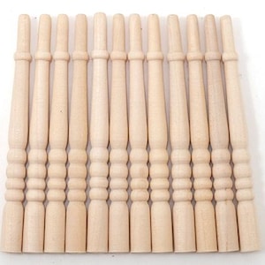 May include: A collection of eleven unfinished wooden spindles, each with a turned design featuring a series of rounded and cylindrical shapes. The spindles are light beige in color and are arranged closely together against a white background.
