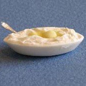 Peut inclure: Un bol ovale miniature blanc rempli d'une substance blanche texturée ressemblant à de la purée ou de la crème, surmonté d'une petite noisette de beurre jaune et doté d'une minuscule cuillère en métal argenté sur le bord gauche.