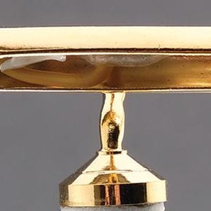 May include: A gold-toned metal display stand featuring a horizontal, hollow, rectangular bar top supported by a central, slender, vertical post that connects to a circular, tiered base. The interior of the bar shows a white plastic clip mechanism.