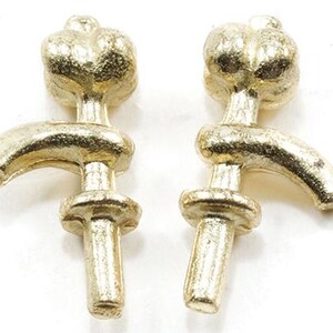May include: A pair of gold-toned metal objects shaped like stylized water faucets. Each piece features a rounded top, a curved spout extending to the side, and a circular disc detail on the vertical stem, all with a textured, matte metallic finish.