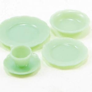 Puede incluir: Un juego de té en miniatura verde jade, que incluye una taza y un platillo, dos platos y un cuenco pequeño. El juego tiene un diseño de borde festoneado. Las piezas están dispuestas sobre una superficie blanca.