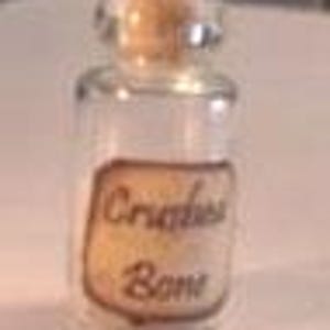 May include: A small, clear glass bottle with a cork stopper. The bottle features a rectangular, aged-looking paper label with dark brown, cursive text that reads Crushed Bone. The bottle is empty and has a cylindrical shape with a narrow neck.