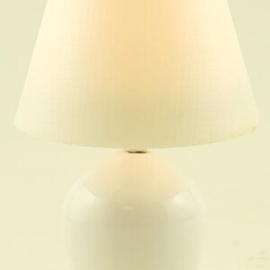 May include: A table lamp featuring a smooth, glossy white ceramic base with a rounded, bulbous shape. The lamp is topped with a tapered, off-white fabric lampshade that emits a soft, warm glow. A thin white power cord extends from the base.