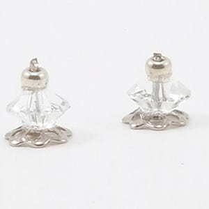 May include: A pair of stud earrings featuring clear, faceted, diamond-shaped crystals. Each crystal is set atop a silver-toned, scalloped metal base and secured with a small, round silver-toned metal post backing at the top of the crystal.