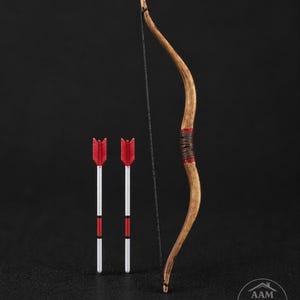 May include: A miniature wooden bow with a drawn string, next to two arrows. The bow is brown with red accents and a wrapped grip. The arrows have red fletching, white shafts, and red and white details.
