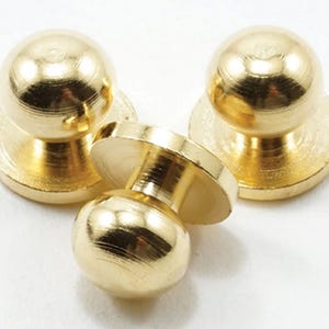 May include: Three gold-colored metal buttons, each featuring a smooth, spherical top attached to a flat, circular base. The buttons have a polished, reflective finish and are arranged in a cluster, showing both the rounded tops and the flat bases.