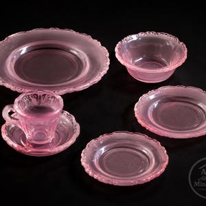 Könnte beinhalten: Eine Sammlung von rosa Miniaturgeschirr aus Glas, darunter ein großer Teller, eine kleine Schüssel, zwei kleinere Teller und eine Teetasse mit Untertasse. Die Ränder der Teller und der Schüssel haben ein gewelltes Design. Das Set wird auf einer schwarzen Oberfläche präsentiert.