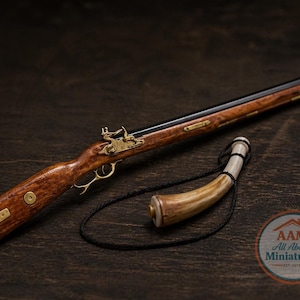 Puede incluir: Una réplica en miniatura de un rifle de chispa con culata de madera y detalles en latón. Un cuerno de pólvora curvado con un cordón negro está junto al rifle. Los objetos están sobre una superficie de madera oscura.