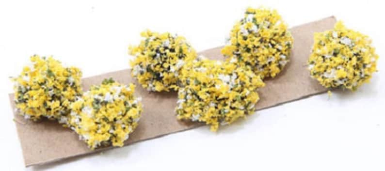 May include: Six small yellow and white flower clusters on a brown paper background. The clusters are made of small, fluffy pieces of material.