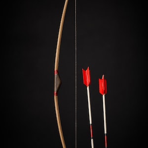 Puede incluir: Un arco de madera en miniatura con una empuñadura marrón oscuro y detalles rojos, acompañado de dos flechas con plumas rojas. El arco está encordado con una cuerda fina y oscura. El fondo es oscuro.