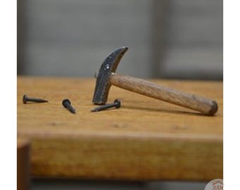 May include: A miniature metal hammer with a wooden handle resting on a wooden surface alongside three small metal nails. The hammer features a curved claw and a dark, textured finish on the head, while the handle is light brown wood.