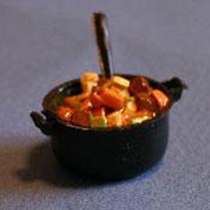May include: A miniature black metal pot with two side handles containing a colorful assortment of small, textured food pieces including orange, yellow, and brown shapes, with a single dark vertical utensil standing upright in the center.