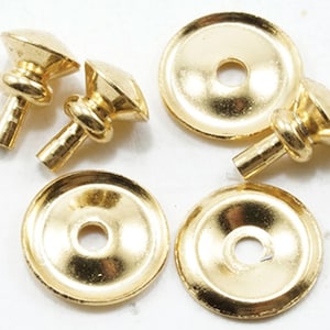 May include: A set of gold-colored metal fasteners consisting of three stud-like components with pointed tops and two flat, circular washers with a central hole. These hardware pieces feature a polished, reflective finish and a smooth, metallic texture.