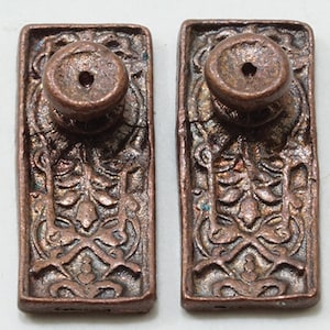 May include: Two antique copper door knobs with ornate floral designs. The knobs are rectangular and have a round, raised center.