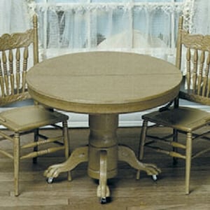 May include: A light brown, round table and two matching chairs. The table has a pedestal base with claw feet and wheels. The chairs have spindle backs and solid seats. The furniture set is made of wood with a distressed finish.