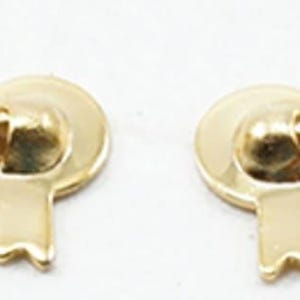 May include: Two matching gold-toned metal drawer pulls featuring a decorative shield-like backplate with a central crest shape and a curved, swinging bail handle. The hardware has a polished, reflective finish and a symmetrical, ornate design.