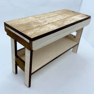 Puede incluir: Una pequeña mesa de madera con una tapa de color marrón claro y una base blanca. La mesa tiene un estante debajo de la tapa.