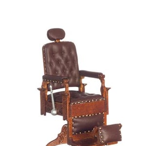 May include: Miniature barber chair with brown leather upholstery and wooden frame. The chair features a headrest, armrests, and a footrest. The chair has a lever on the side and is designed with a classic, vintage aesthetic.