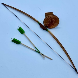 Könnte beinhalten: Ein Miniatur-Holzbogen mit dunkler Schnur, braunen und grünen Akzenten und zwei Pfeilen mit grünen Federn. Eine Münze dient zum Größenvergleich. Der Bogen ist ein handgefertigter Artikel.