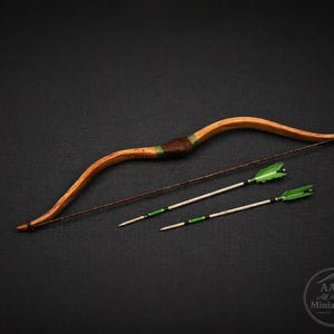 Puede incluir: Un arco de madera en miniatura con una empuñadura marrón y dos flechas con plumas verdes. El arco es de madera marrón claro y tiene una cuerda marrón oscuro. Las flechas son blancas con detalles verdes y negros.