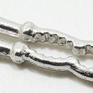 May include: A silver-colored metal nutcracker featuring two elongated handles connected by a central hinge, with a textured, scalloped inner gripping area designed for cracking shells, presented in a simple, functional, and metallic finish.