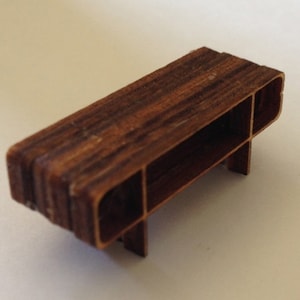 May include: A miniature wooden table with a brown wood grain finish. The table has a rectangular top and three legs. The table is made of layered wood.